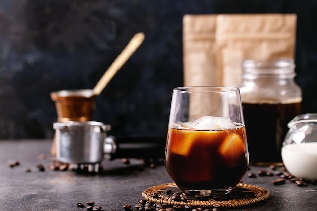 Glass of ice coffee with ice cubes served with cream, copper cezve and coffee beans over dark wall