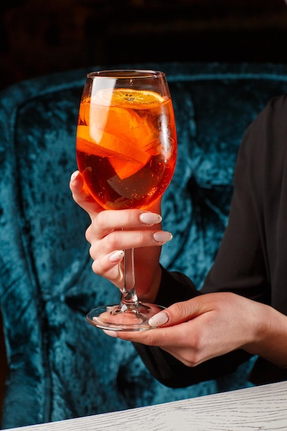 Glass of ice cold Aperol spritz cocktail served in a wine glass in female hands decorated with slices of orange placed on a black background