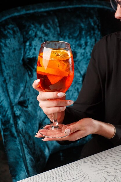 Glass of ice cold Aperol spritz cocktail served in a wine glass in female hands decorated with slices of orange placed on a black background