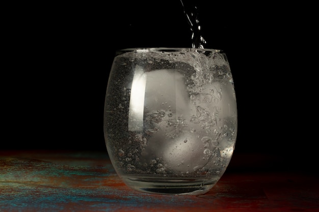 Bicchiere di acqua ghiacciata che viene riempito con acqua frizzante fredda su uno sfondo scuro e superficie rustica.