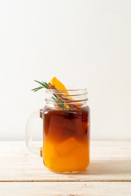 A glass of iced americano black coffee and layer of orange and lemon juice decorated with rosemary and cinnamon on wooden table