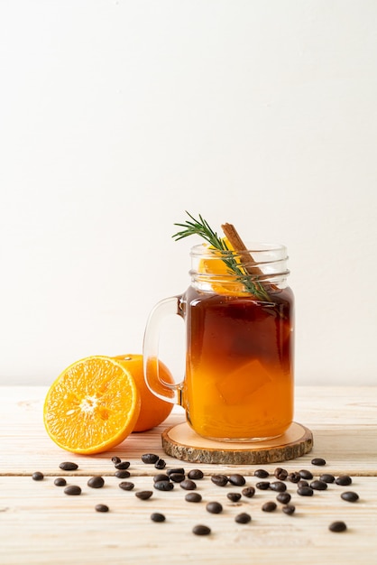 A glass of iced americano black coffee and layer of orange and lemon juice decorated with rosemary and cinnamon on wooden table