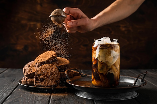 Un bicchiere di caffè freddo con biscotti al latte e cioccolato su un piatto, spolverando i biscotti con cacao in polvere