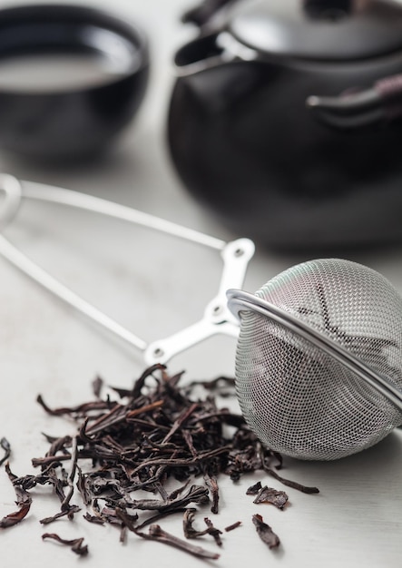 Glass jar of black loose organic tea with teapot and cup with tea strainer infuser on light background