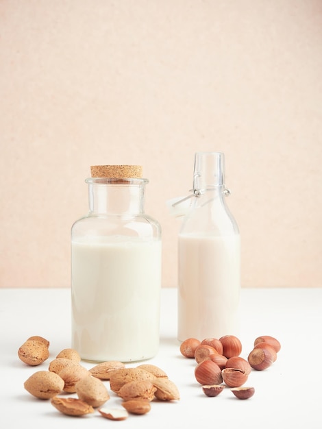 Glass jar and bottle filled with vegetable nut drink placed on white table near heap of almonds and hazelnuts near pink wall Vegan dairy free milk