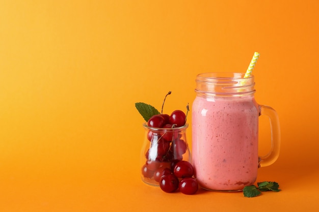 Glass jar of cherry smoothie and ingredients on orange background