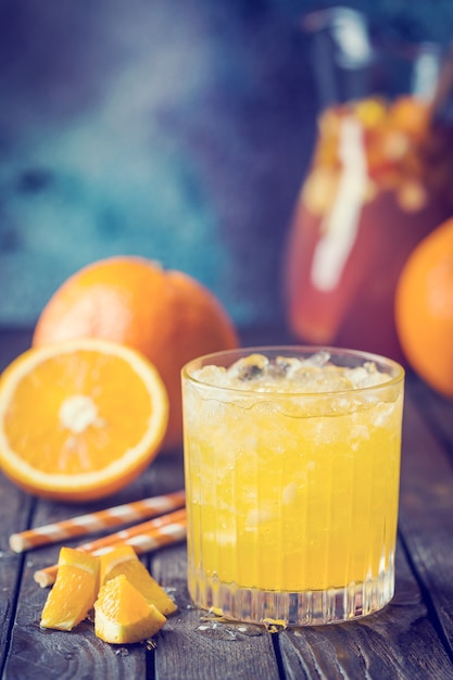 Glass jar of fresh orange juice with ice and fresh fruits on dark table