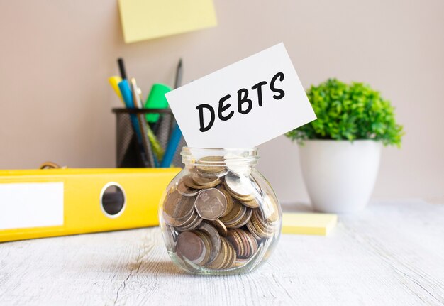 The glass jar is filled with coins. On the bank there is a card with the inscription DEBTS. Everything is located on the office desk. Financial concept.