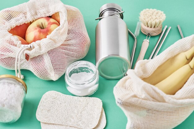 Glass jar, metal straws, aluminum bottle, wooden cleaning brush and fruits in shopping bag, top view