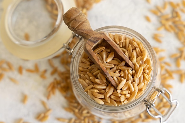 Glass jar of raw dry rye grain with a wooden spoon on white table close up