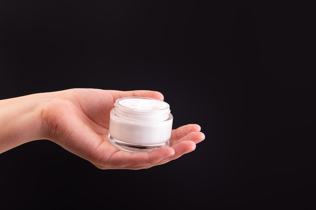 Glass jar of white moisturized cream in a female hand on black background Body care lotion Simple