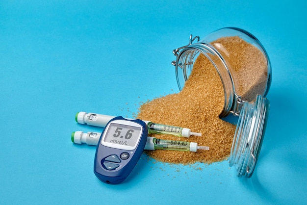 Glass jar with brown cane sugar insulin syringes and a glucometer on a blue background