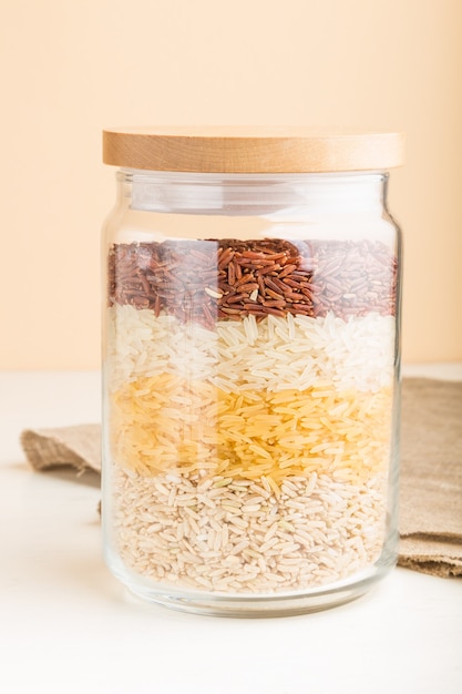 Glass jar with different kinds of rice poured in layers on white and orange surface. side view, close up