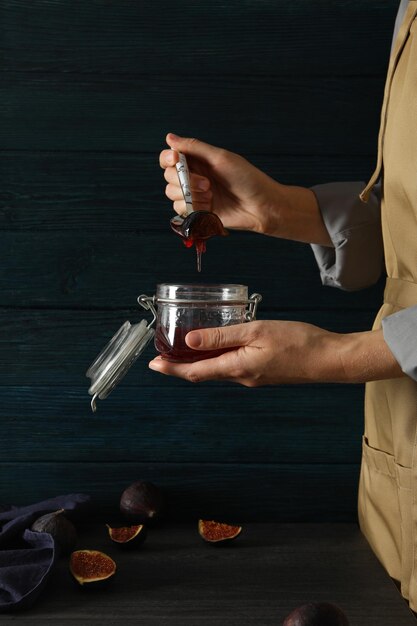 Vaso di vetro con marmellata di fichi e cucchiaio nelle mani femminili asciugamano e fichi tagliati su tavolo su sfondo di legno