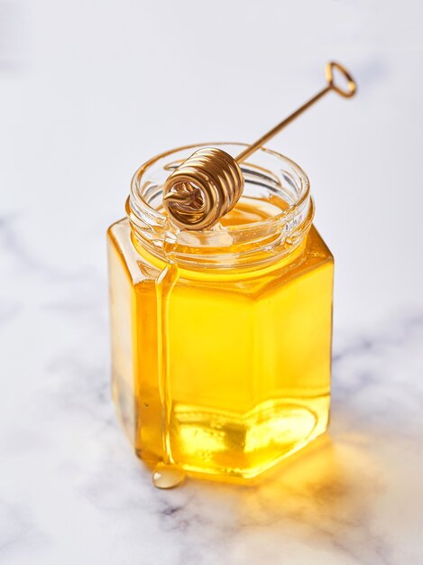 Glass jar with floral liquid honey and metal honey spoon on light marble background. Alternative sugar substitute, cold remedy and body strengthening, superfood