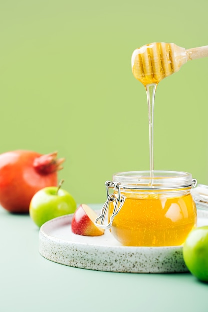 Glass jar with fresh honey, honey spoon, apple and pomegranate on a bright green background. Creative Concept Jewish New Year Happy holiday Rosh Hashanah. layout of traditional symbols.View from above