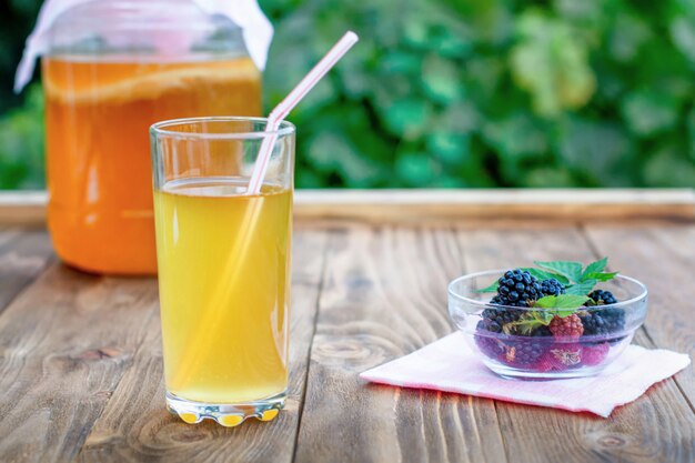 Glass jar with kombucha, a poured glass with kombucha and a cup with blackberry in a summer garden.