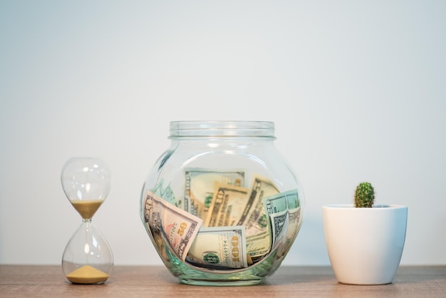 Glass jar with money on the table sand clock and cactus nearby