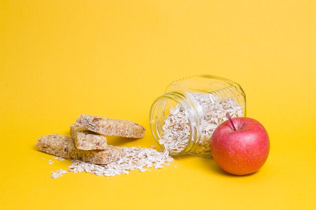 Barattolo di vetro con farina d'avena, una mela e diverse barrette proteiche per uno spuntino su una superficie gialla