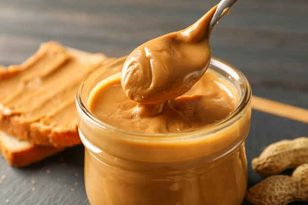 Glass jar with peanut butter and spoon, peanut, peanut butter sandwich and cutting board on wooden table