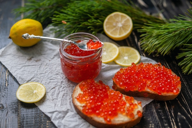 Glass jar with red caviar next to lemons and pine needles on a black wooden background