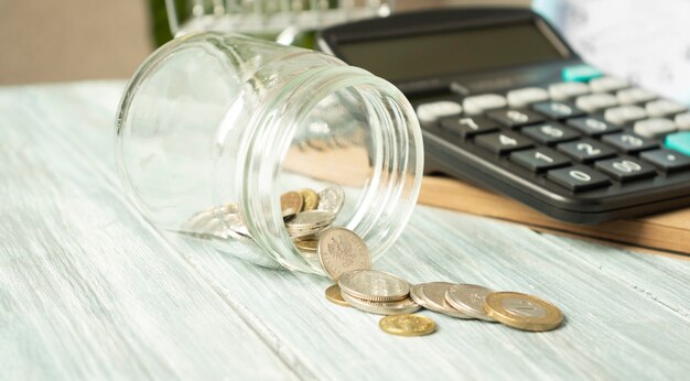 Glass jar with scattered coins and a calculator on a wooden table.