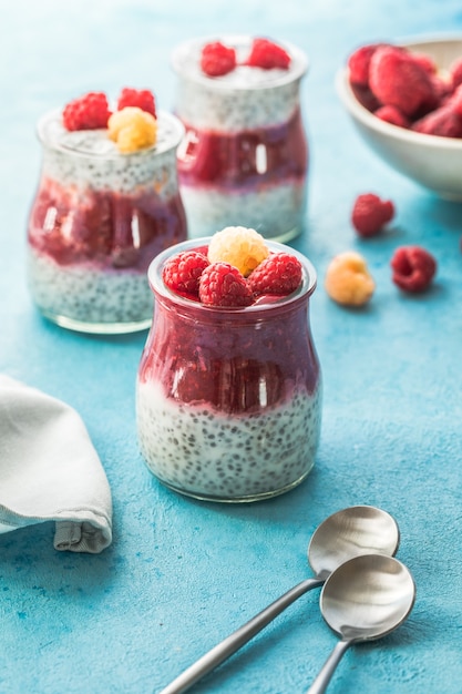 Glass jars with chia pudding with raspberry and jam. Bowl of raspberry berry on the blue background.