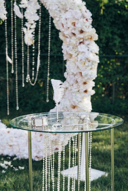 Glass jewelry box next to a pen for writing on a glass table decorated with glass beads against a white flower arch