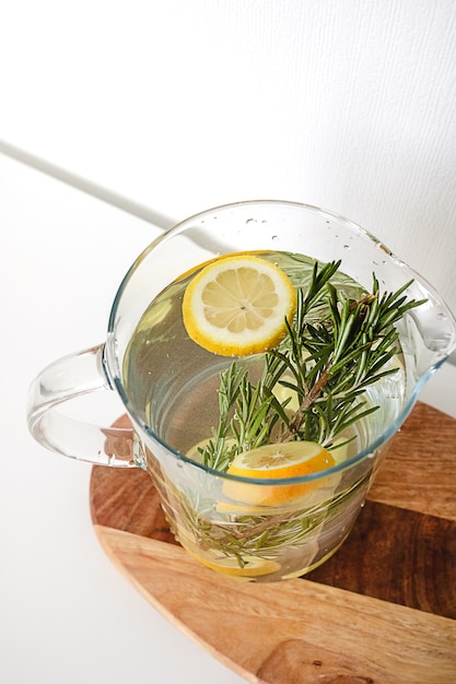 A glass jug of water with lemon and rosemary on a wooden cutting board on a white background