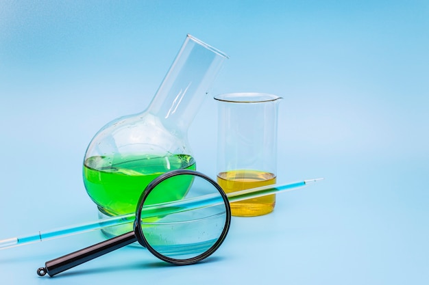 Glass laboratory flask and beaker in a cup with a green and yellow solution with a magnifying glass on a gentle blue medical background. The concept of medical and chemical analyzes. copy space.