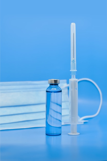 Glass medical ampoules, syringe and protective mask on blue background. Pharmaceuticals, vaccination