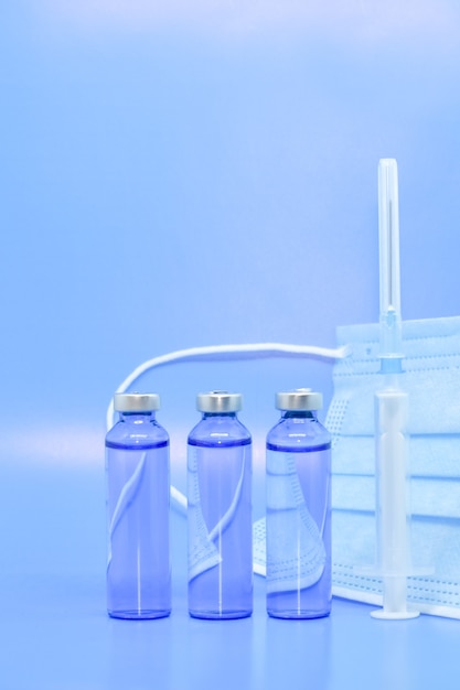 Glass medical ampoules, syringe and protective mask on blue background. Pharmaceuticals, vaccination