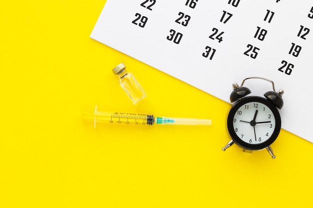 黄色の背景に液体と注射器、カレンダーと目覚まし時計が付いているガラスの薬のバイアル。医療ワクチン接種スケジュール。健康の概念。フラットレイ、コピースペース付きの上面図。