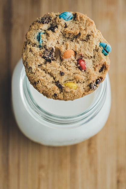 Glass of milk and homemade sugar cookies with colorful chocolate candies on a wooden board. Copy space. Top view.