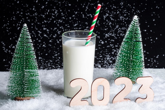 A glass of milk with a straw on a snowy background among toy Christmas trees and wooden numbers 2023