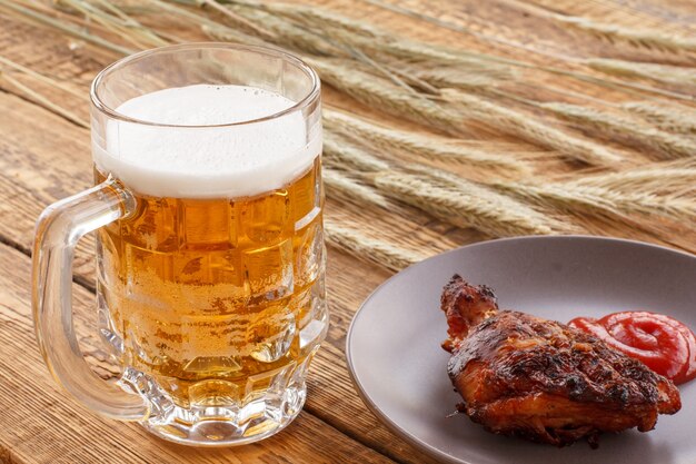 Glass mug of beer, plate with grilled chicken and tomato sauce, ears of barley on a wooden background.