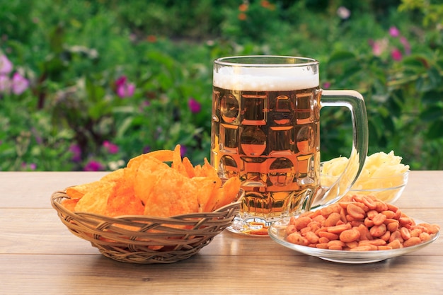 Glass mug of beer on wooden table with potato chips in wicker basket, peanuts and dried squid in bowls