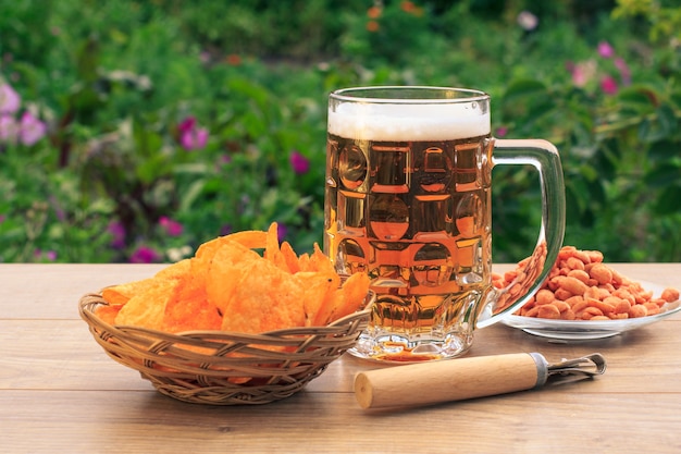 Glass mug of beer on wooden table with potato chips in wicker basket, peanuts in plate, opener