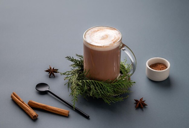 Photo glass mug of hot chocolate with whipped cream and cinnamon stick on a stand with pine branches on a gray background with anise star spoon and cocoa powder
