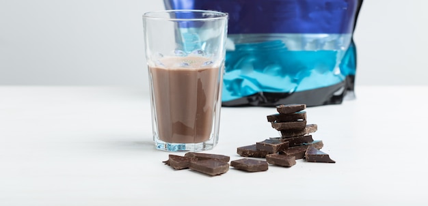 Glass mug with a protein chocolate shake stands against the\
background of a packet of protein on a white table. sports\
nutrition concept.