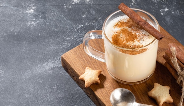 Glass mug with traditional Christmas drink homemade creamy eggnog with cinnamon and heart shape cookies on wooden kitchen board on grey. Typical holiday dessert. Close up. Copy space.