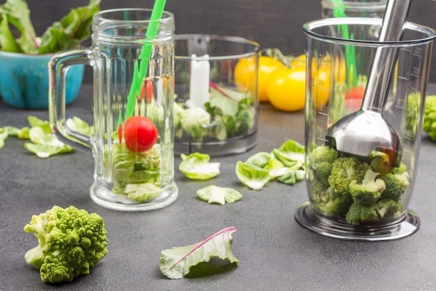 Glass mugs with broccoli, tomato and green straws. Blender bowl with Metal shredder