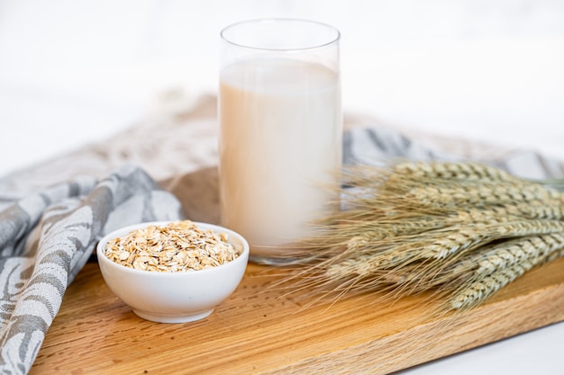 Glass of oat milk with oat flakes on white wooden background