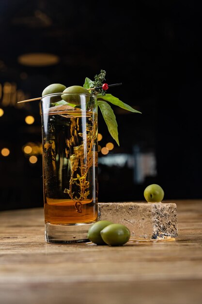 A glass of olives sits on a table with a bottle of alcohol on the table.