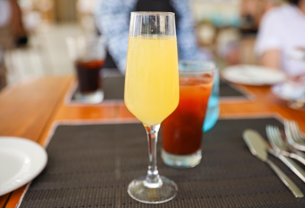 A glass of orange juice sits on a table with a drink in the background.