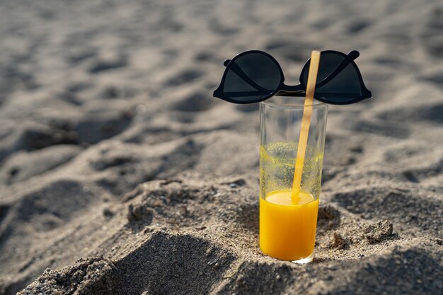 a glass of orange juice with a straw stands on the sand against the backdrop of the sea