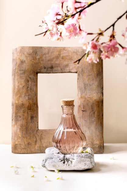 Glass pink transparent bottle of perfume with cork standing on textured stone on white marble table with white flowers and blossom spring branch Perfume making concept with place for design label