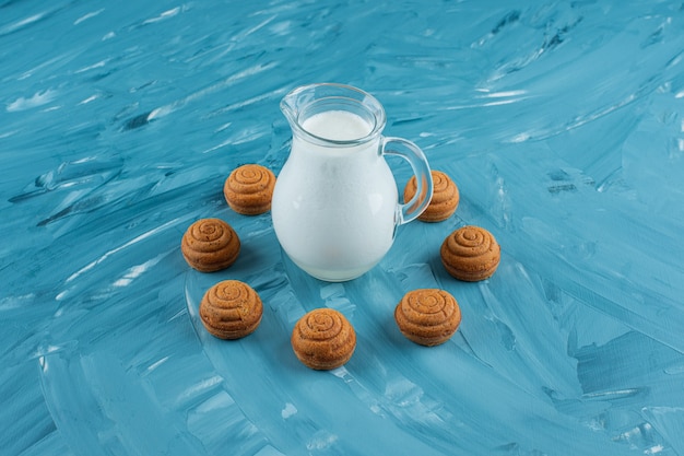 A glass pitcher of fresh milk with sweet round cookies on a blue surface