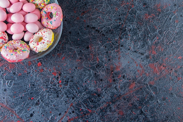 Glass plate of colorful delicious donuts and pink candies on dark surface. 