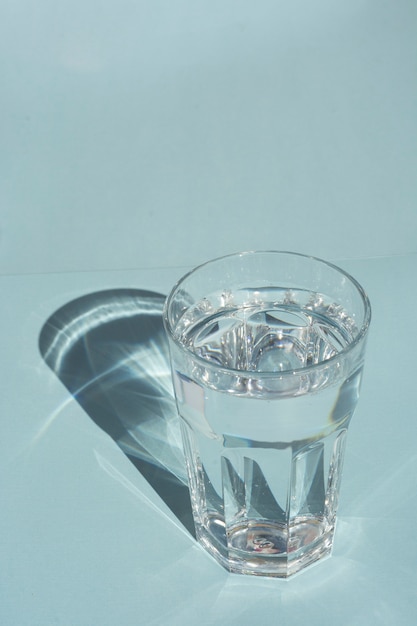 A glass of pure water under sunlight with deep stylish shadows on blue  copyspace